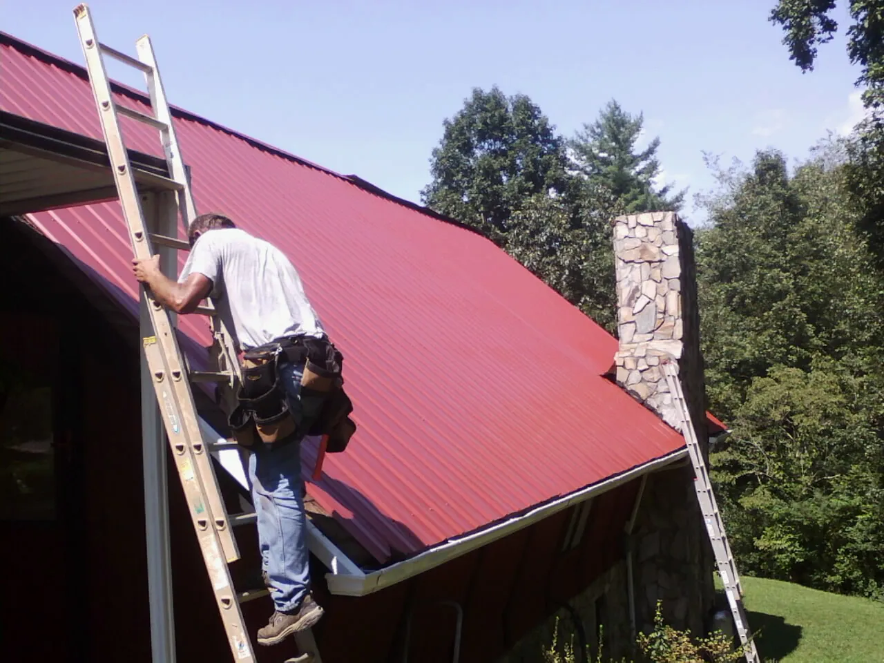 Roofer on a ladder beside a red metal roof and stone chimney — Soffit & Fascia Repair by Roofing Brothers in Teasdale